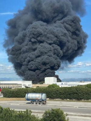 A large, thick cloud of black smoke rises into the sky from a fire behind industrial buildings, with a tanker truck parked on the road in the foreground.